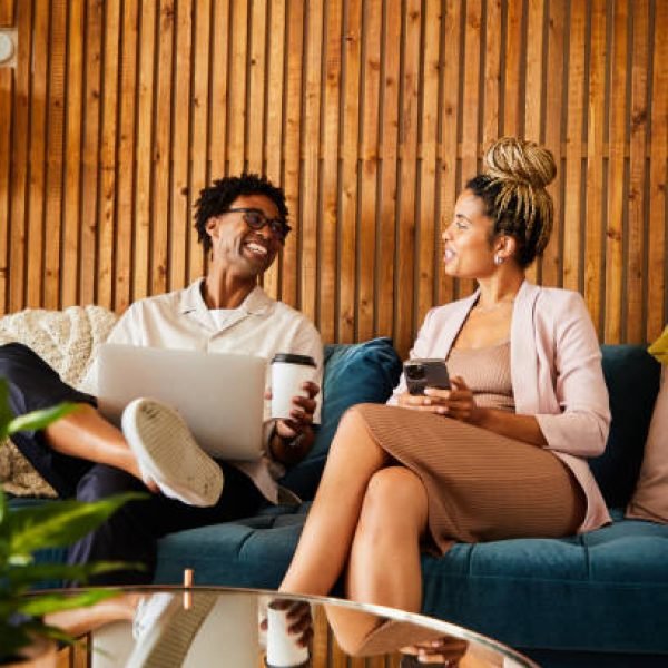 Two laughing young African American businesspeople talking while using different devices while sitting on an office sofa during a break